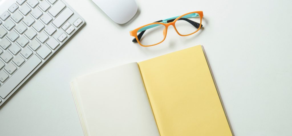 A photo of glasses on a desk with a keyboard and open notebook