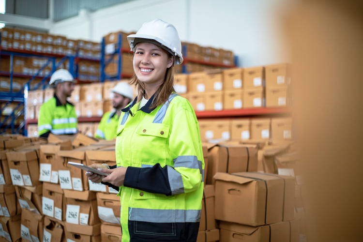 A happy female staff in the warehouse, with proper work gear and ipad in her hand updating the logs.